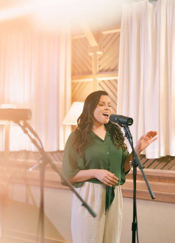 Rayre Mota cantando ao microfone em um estúdio, com luz quente e cortinas ao fundo
